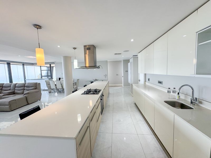 Modern open-plan kitchen featuring a long island with gas cooktop, sleek gloss cabinetry, stone countertops, and integrated appliances, flowing into the living and dining area.