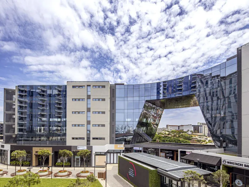 A modern apartment complex featuring a striking glass facade, architectural bridge design, and a well-maintained landscaped courtyard with commercial retail spaces at ground level.