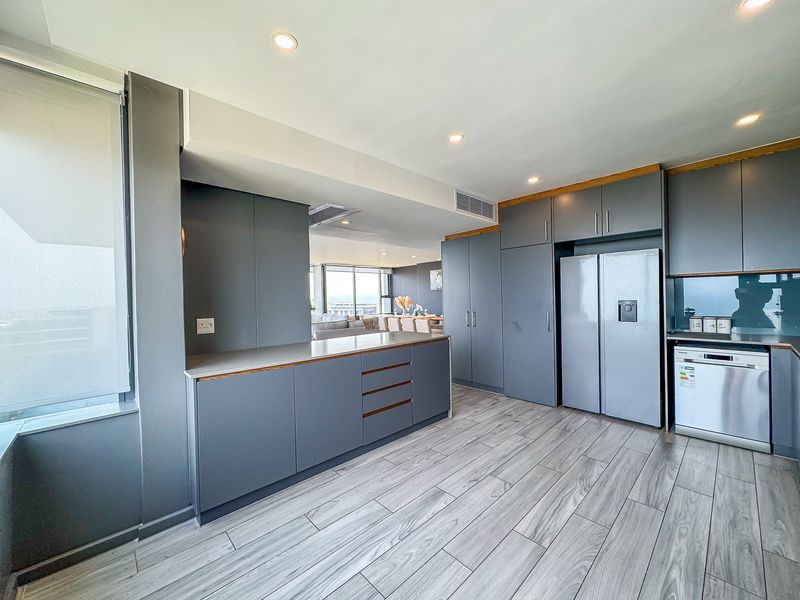 Modern kitchen featuring sleek grey cabinetry, stone countertops, integrated dishwasher, and wood-look tile flooring, seamlessly flowing into the adjacent living area.