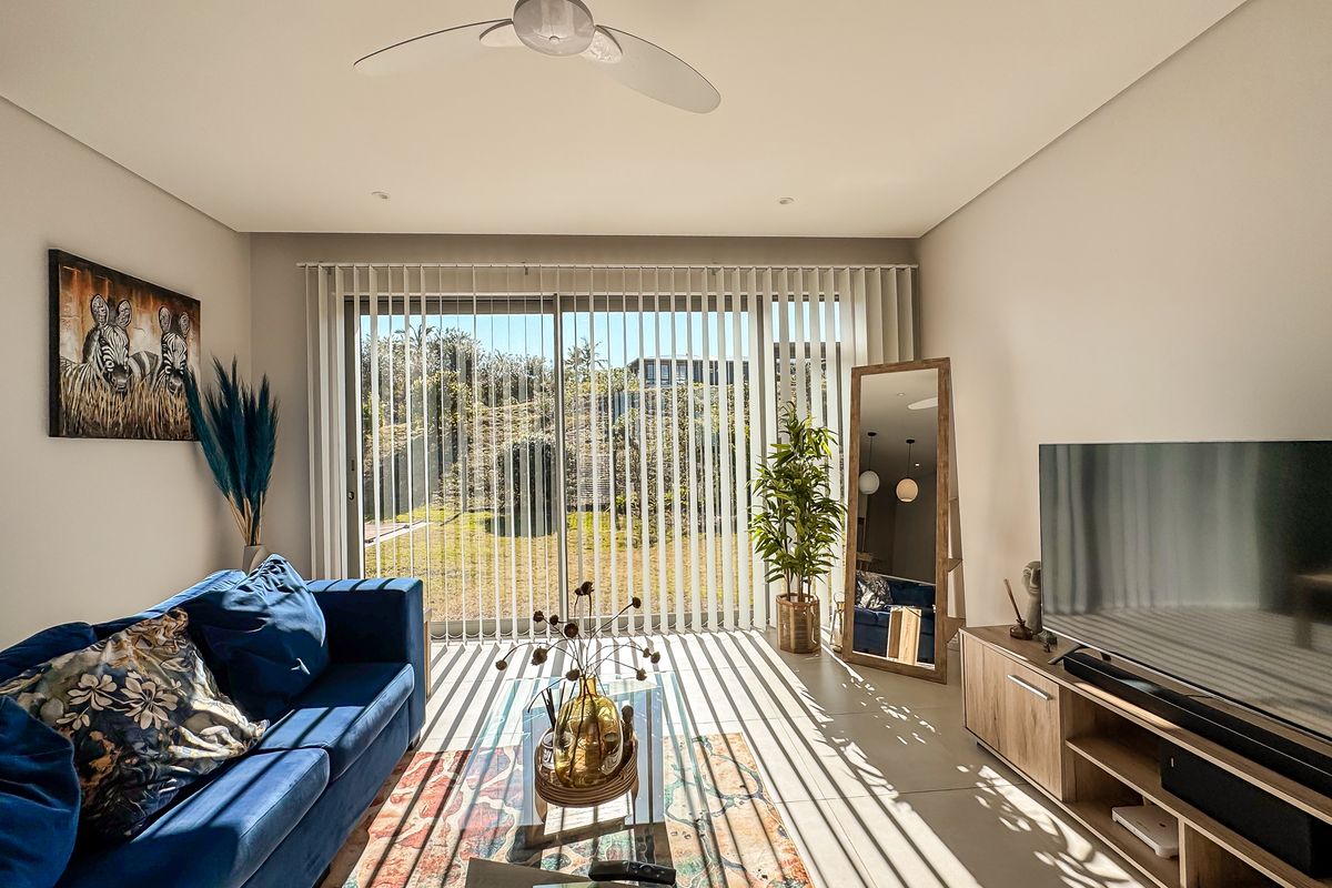 A bright living room featuring tiled flooring, a ceiling fan, and large sliding glass doors with vertical blinds providing access to the garden.