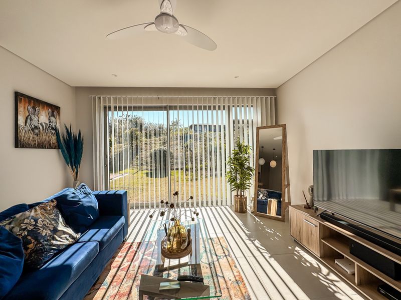 A bright living room featuring tiled flooring, a ceiling fan, and large sliding glass doors with vertical blinds providing access to the garden.