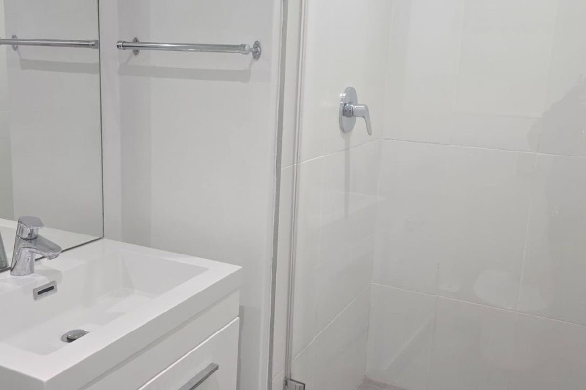 A clean, modern bathroom featuring a white vanity with basin, chrome mixer tap, towel rail, and a glass-enclosed shower with white wall tiles.
