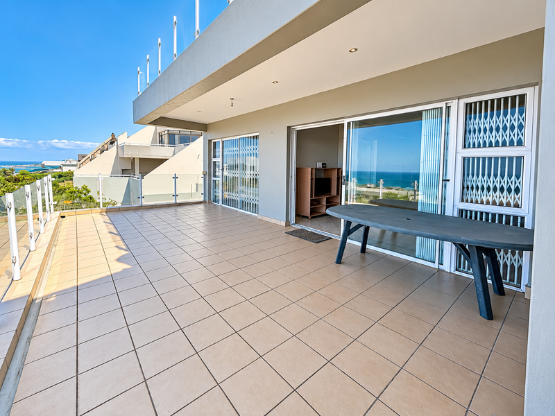 Spacious covered balcony featuring tiled flooring, glass balustrades, and sliding door access, offering an expansive outdoor living area with scenic coastal views.