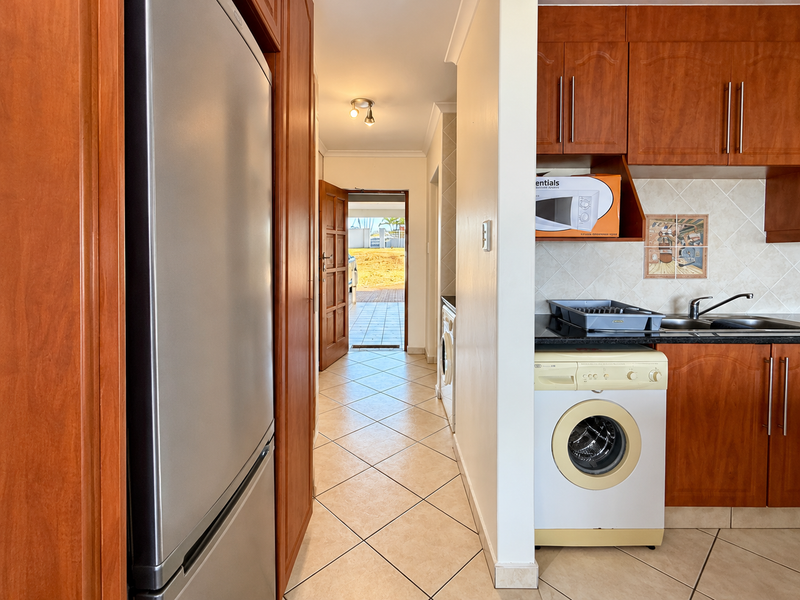 A functional kitchen area featuring wood-finish cabinetry, tiled backsplash, and integrated laundry plumbing, leading through to an exterior entrance.