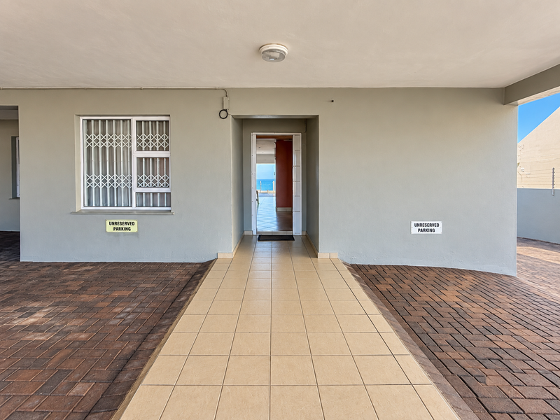 Covered entrance area featuring a tiled walkway, paved driveway, and security gate, providing access to the apartment with a view of the ocean beyond.