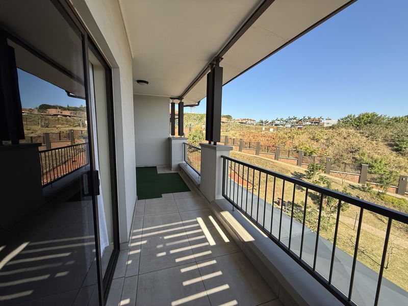 A spacious covered balcony featuring tiled flooring, a metal balustrade, and sliding glass door access, offering pleasant views of the surrounding landscape.