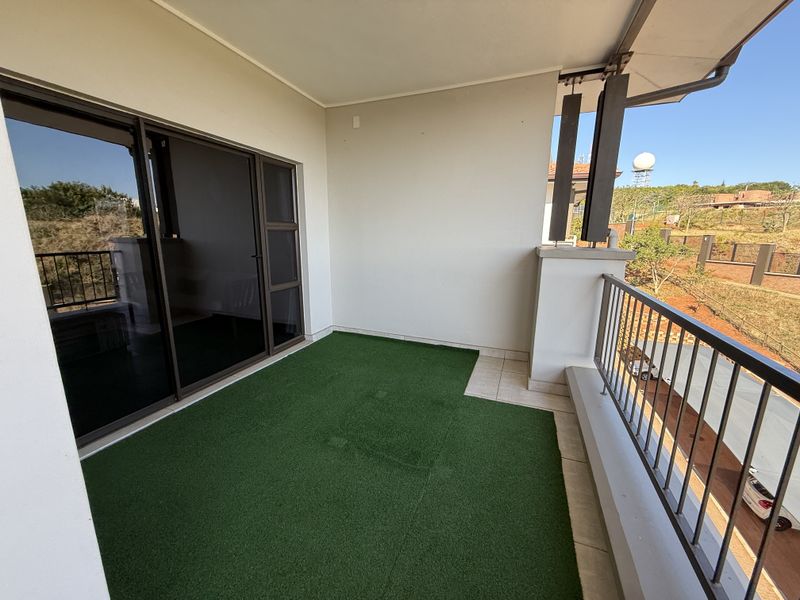 A covered balcony featuring artificial grass, tiled sections, and a metal balustrade, offering an open outdoor space accessible via sliding glass doors.