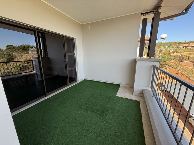 A clean, covered balcony featuring tiled flooring with artificial grass, a metal balustrade, and large sliding glass doors leading into the apartment.