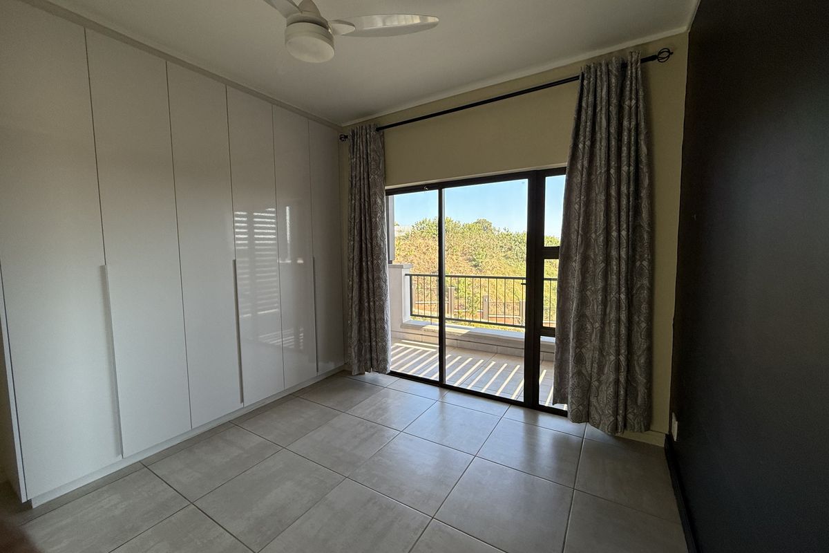 A bright, empty bedroom featuring modern built-in cupboards, tiled flooring, a ceiling fan, and a sliding glass door leading to a private balcony.