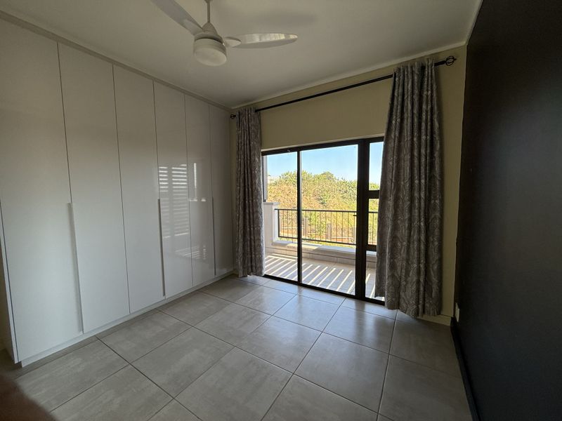 A bright, empty bedroom featuring modern built-in cupboards, tiled flooring, a ceiling fan, and a sliding glass door leading to a private balcony.