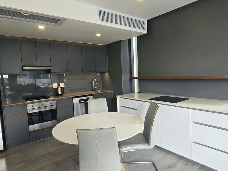 Modern open-plan kitchen and dining area featuring sleek dark cabinetry, integrated appliances, stone countertops, and ducted air conditioning in a contemporary apartment setting.