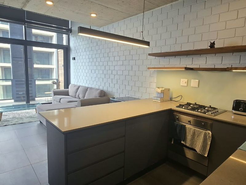 Modern open-plan apartment featuring a sleek kitchen with stone countertops, gas hob, and exposed concrete ceilings, flowing into a bright living area.