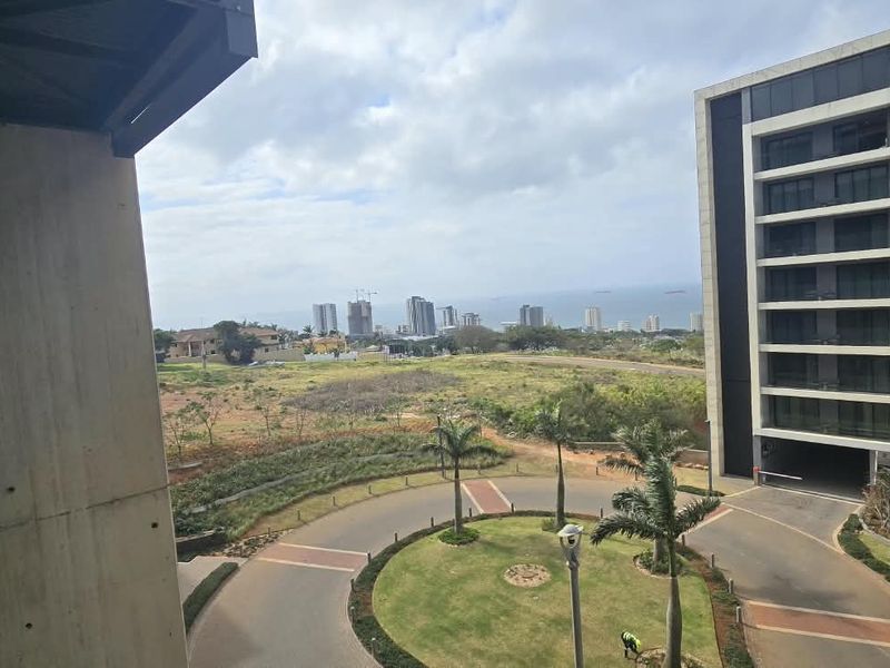 Elevated view from a balcony overlooking a landscaped circular driveway, surrounding apartment complex, and distant coastal cityscape.