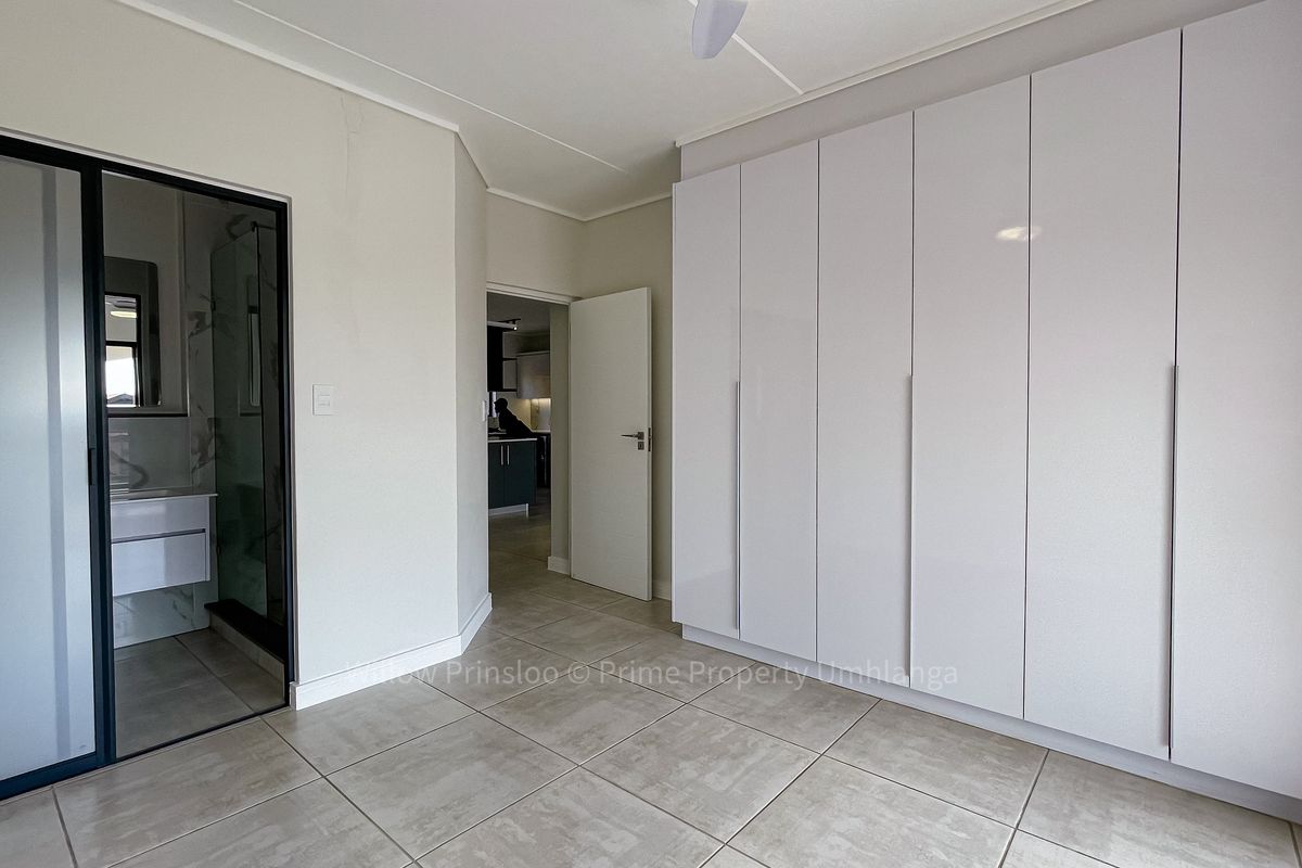 A bright, modern bedroom featuring sleek white built-in cupboards, neutral tiled flooring, a ceiling fan, and direct access to an en-suite bathroom.