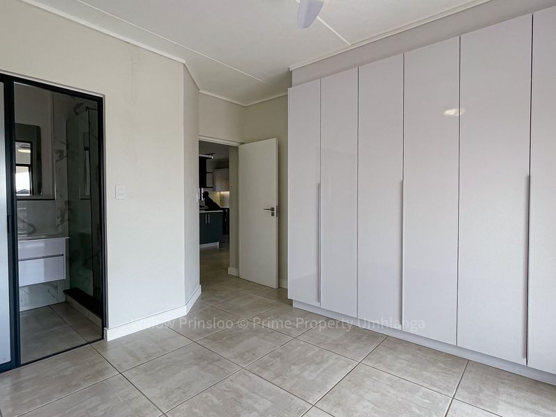 A bright, modern bedroom featuring sleek white built-in cupboards, neutral tiled flooring, a ceiling fan, and direct access to an en-suite bathroom.