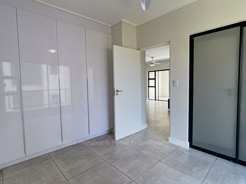 A bright, empty bedroom featuring modern high-gloss built-in cupboards, neutral tiled flooring, and a sleek sliding door leading to an adjacent living space.