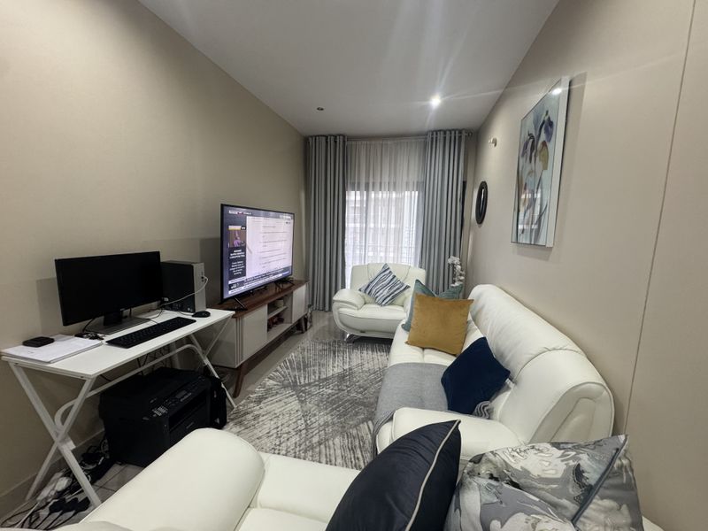 A well-maintained living room featuring neutral wall tones, tiled flooring, and large windows with floor-to-ceiling curtains, providing a comfortable and functional living space.