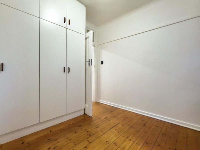 A bright, empty bedroom featuring polished timber flooring, white walls with a picture rail, and ample built-in cupboard storage.