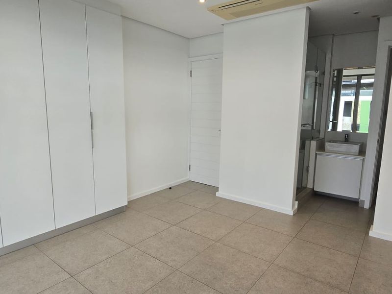A clean, modern bedroom featuring neutral tiled flooring, ample built-in cupboard space, and an integrated en-suite bathroom with a vanity basin.