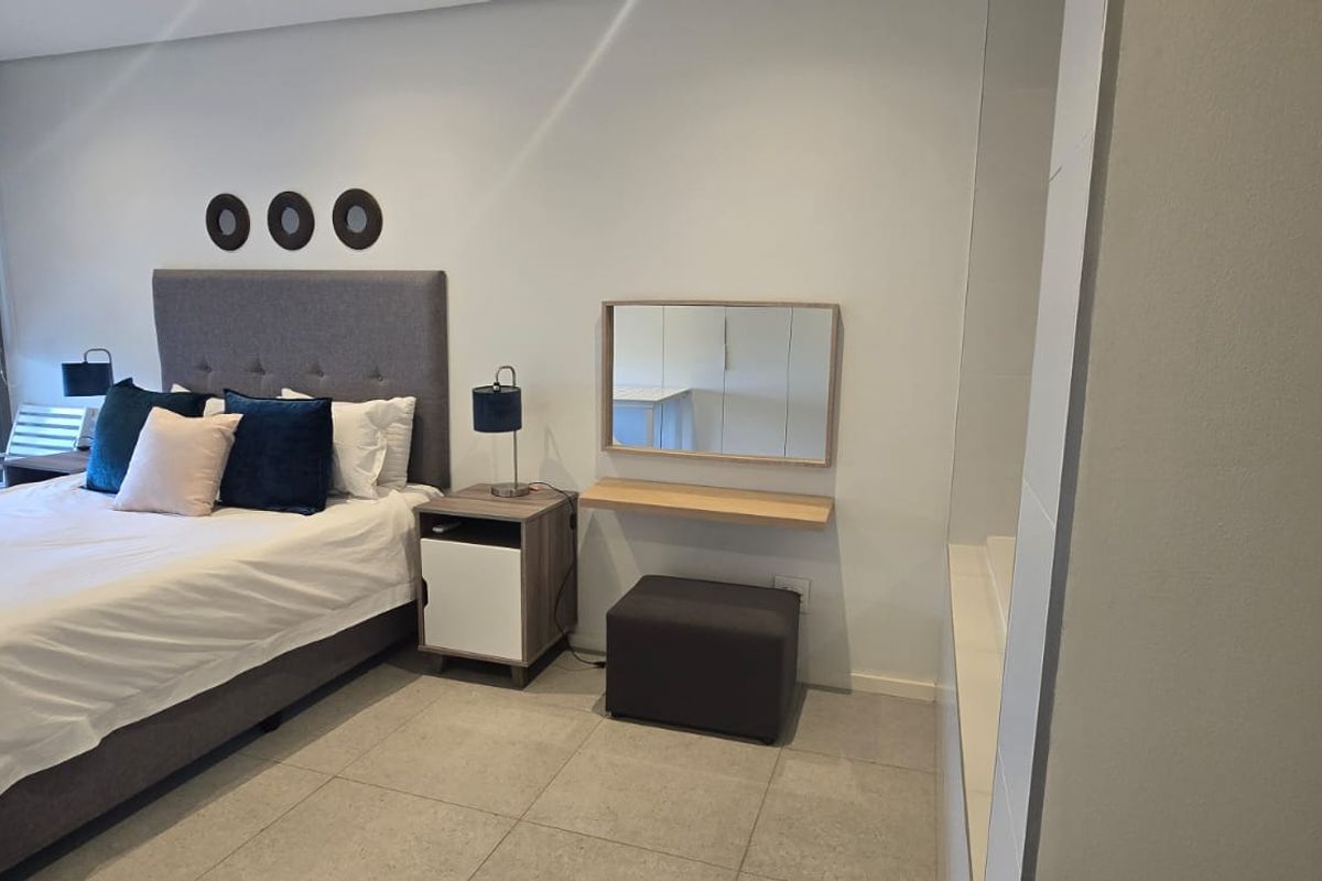 A modern bedroom featuring neutral tiled flooring, a wall-mounted mirror with a floating shelf, and large windows with roller blinds.