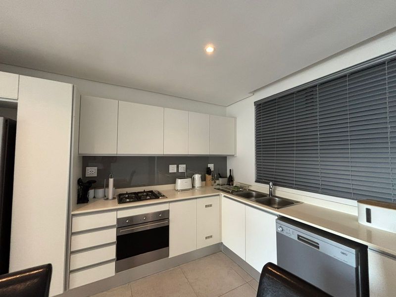 Modern kitchen featuring white cabinetry, gas hob, electric oven, stainless steel sink, and sleek glass splashback with neutral tiled flooring.