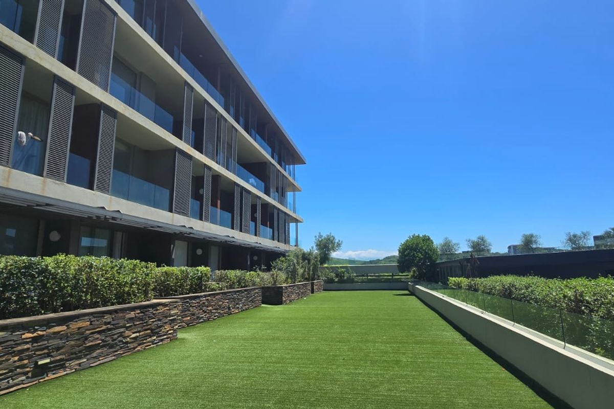 A well-maintained rear garden area featuring artificial turf, stone retaining walls, and glass balustrades, set against a modern multi-story apartment building.
