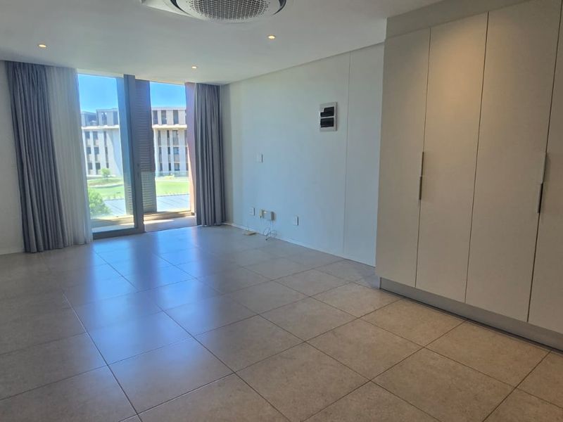 A bright, empty bedroom featuring neutral tiled flooring, ample built-in cupboard space, and a sliding glass door leading to an outdoor area.
