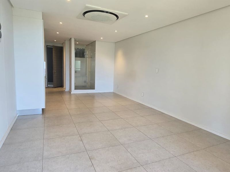 A bright, open-plan living area featuring neutral tiled flooring, recessed ceiling lighting, and a modern ducted air conditioning system.
