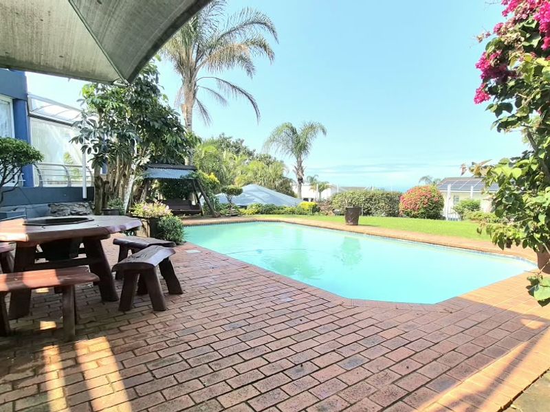 Outdoor pool area featuring brick paving, a shade sail, and a well-maintained garden setting.