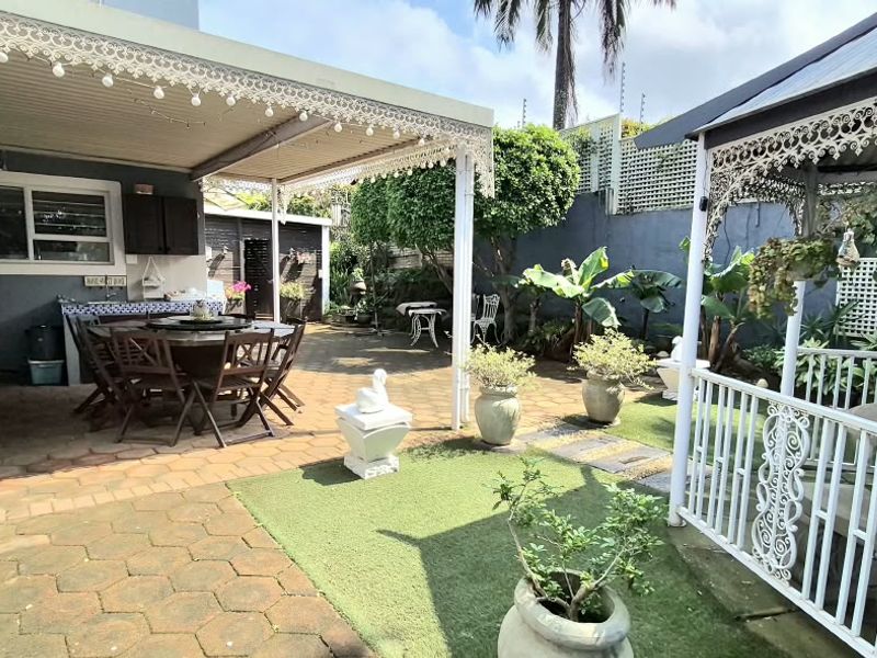 A spacious outdoor entertainment area featuring a covered patio, paved flooring, artificial grass sections, and a decorative wrought iron gazebo.