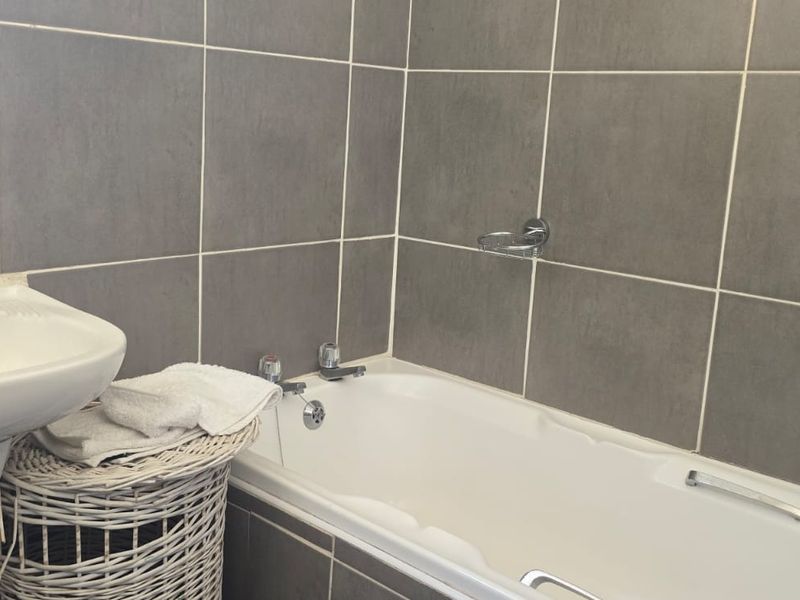 A functional bathroom featuring grey wall tiles, a white pedestal washbasin, and a bathtub with chrome fixtures.