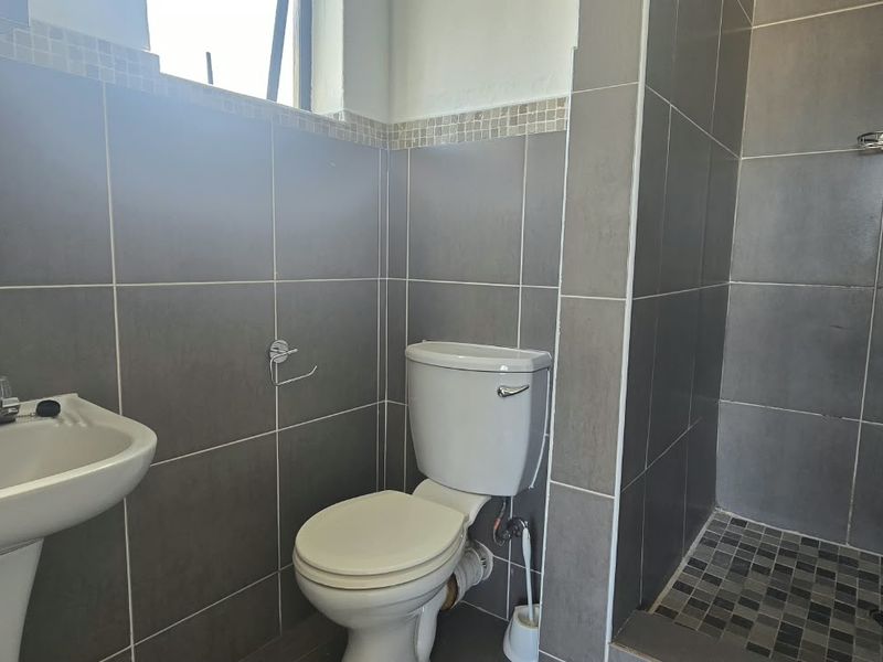 A functional bathroom featuring a pedestal washbasin, toilet, and a walk-in shower with mosaic floor tiling and grey wall tiles.