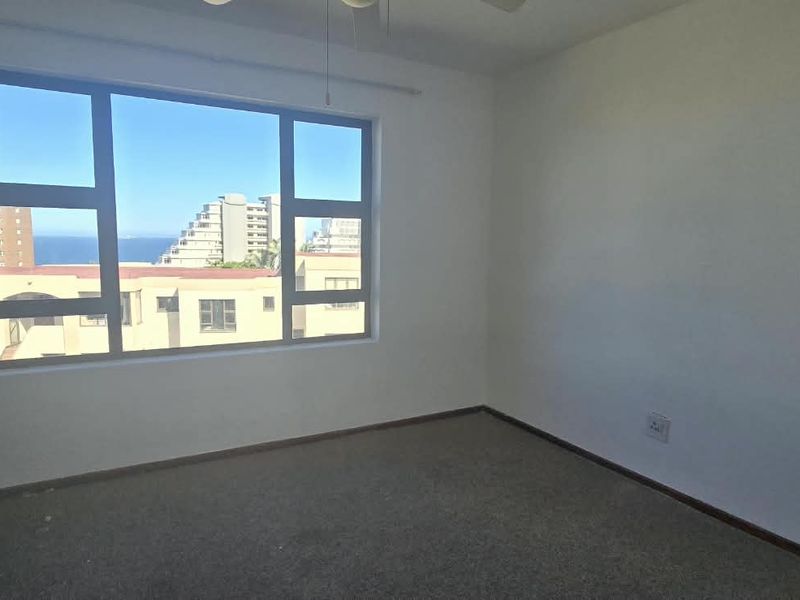 A bright, empty bedroom featuring a built-in wardrobe, ceiling fan, and large windows offering an urban and coastal view.