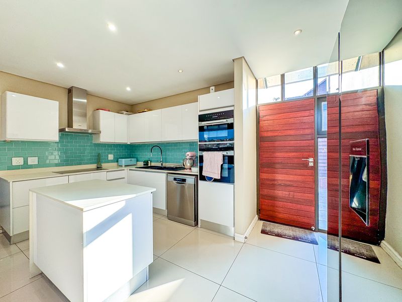 Modern kitchen featuring white cabinetry, a teal subway tile backsplash, built-in oven, kitchen island, and direct access to a timber front entrance door.