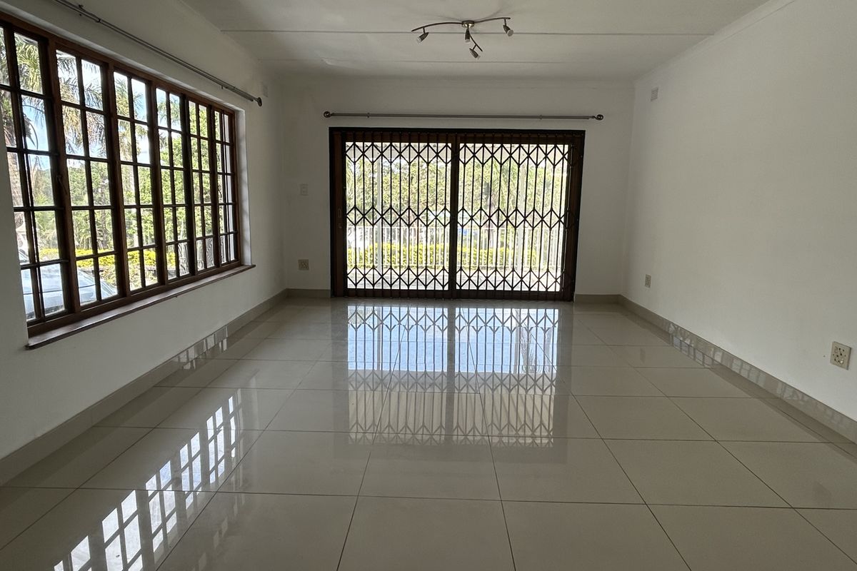 A bright, empty living room featuring large windows, tiled flooring, and a sliding glass door with a security gate leading to the exterior.