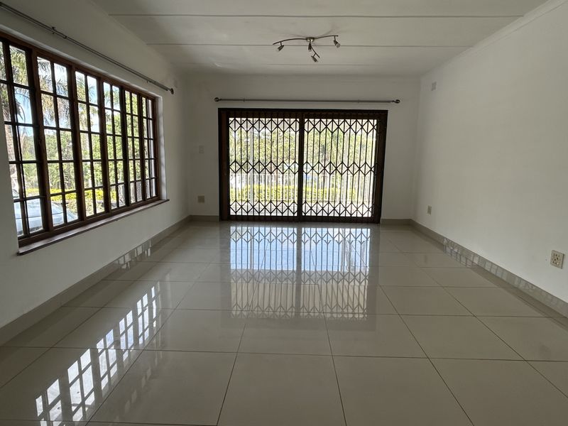 A bright, empty living room featuring large windows, tiled flooring, and a sliding glass door with a security gate leading to the exterior.