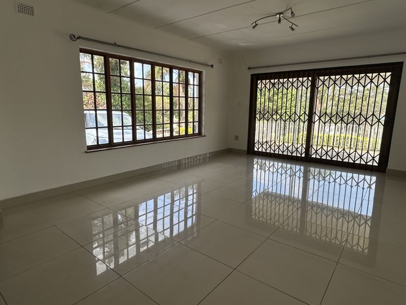 A bright, empty living room featuring polished tiled flooring, large wooden-framed windows, and a sliding glass door with a security gate.