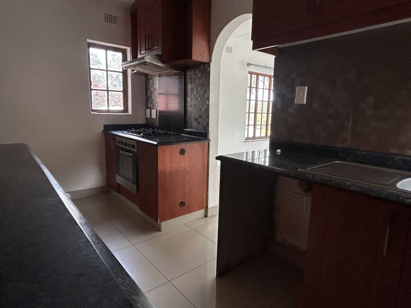 A functional kitchen featuring dark wood cabinetry, granite countertops, built-in oven, gas hob, and tiled flooring, with an open archway leading to adjacent space.