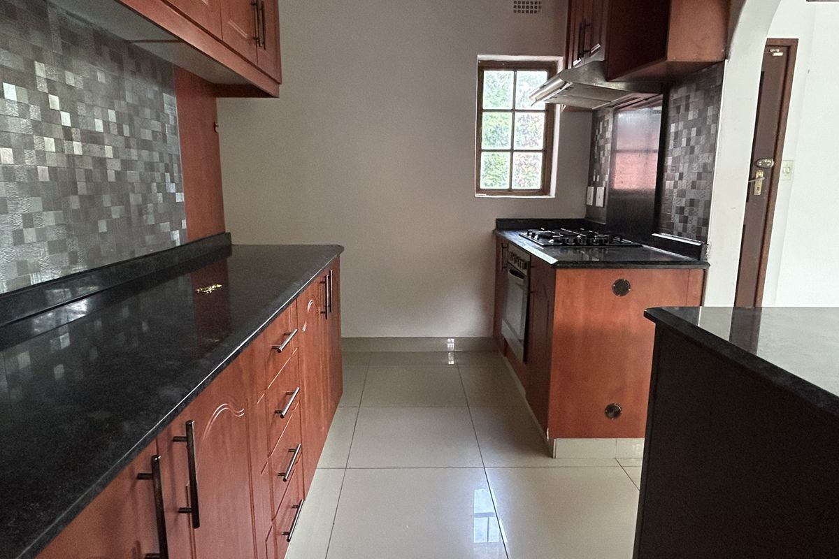 A functional kitchen featuring wooden cabinetry, granite countertops, a gas hob, built-in oven, and mosaic tile backsplash with neutral floor tiling.