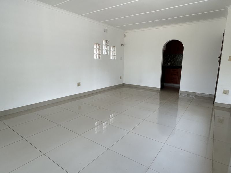 An empty, open-plan living area featuring neutral porcelain floor tiles, a unique glass block window, and an arched entryway leading to the kitchen.