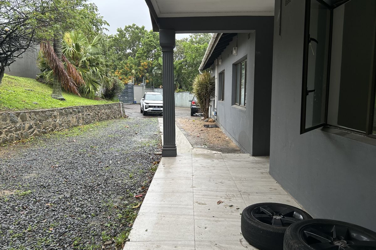 A covered patio area featuring tiled flooring and a support column, overlooking a gravel side yard with a stone retaining wall.