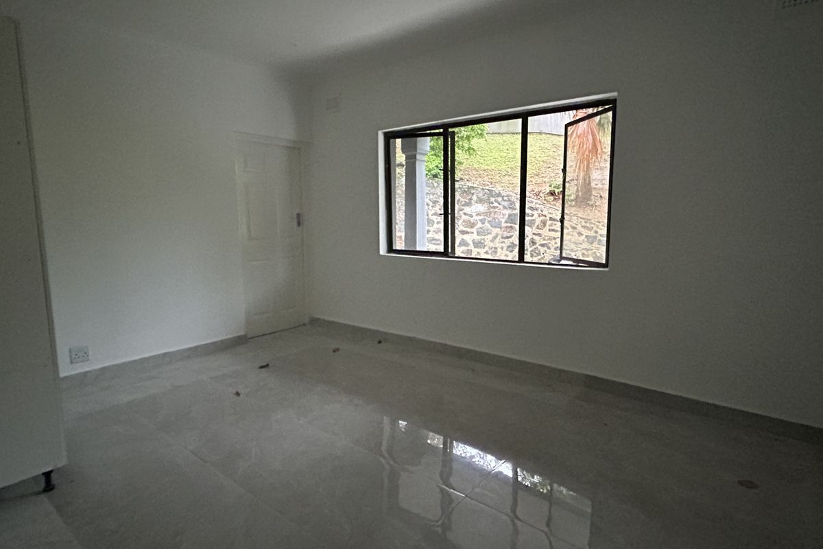 An empty bedroom featuring neutral tiled flooring, white walls, and a large window providing natural light and a view of the exterior stone retaining wall.