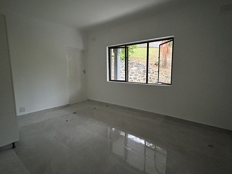 An empty bedroom featuring neutral tiled flooring, white walls, and a large window providing natural light and a view of the exterior stone retaining wall.