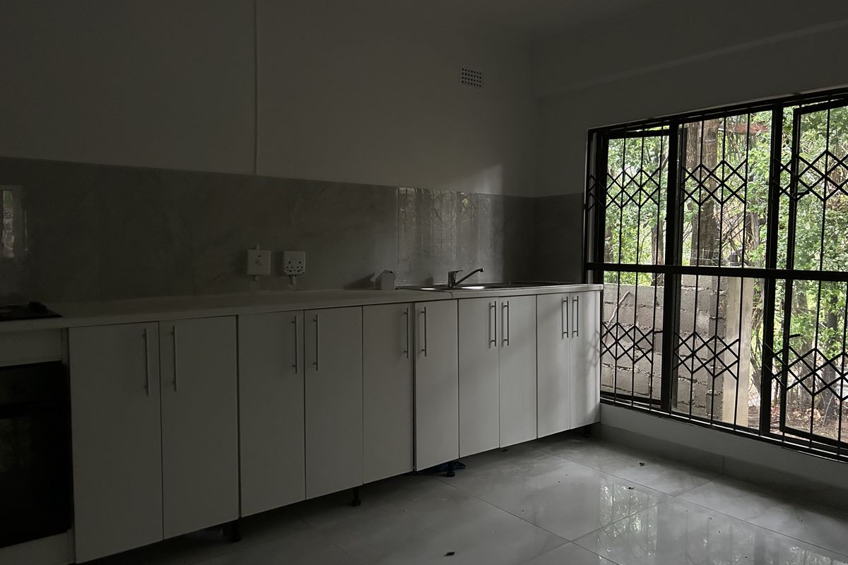 A clean, empty kitchen featuring white built-in cabinetry, a stainless steel sink, tiled splashback, and large windows with security gates.