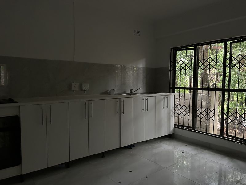 A clean, empty kitchen featuring white built-in cabinetry, a stainless steel sink, tiled splashback, and large windows with security gates.