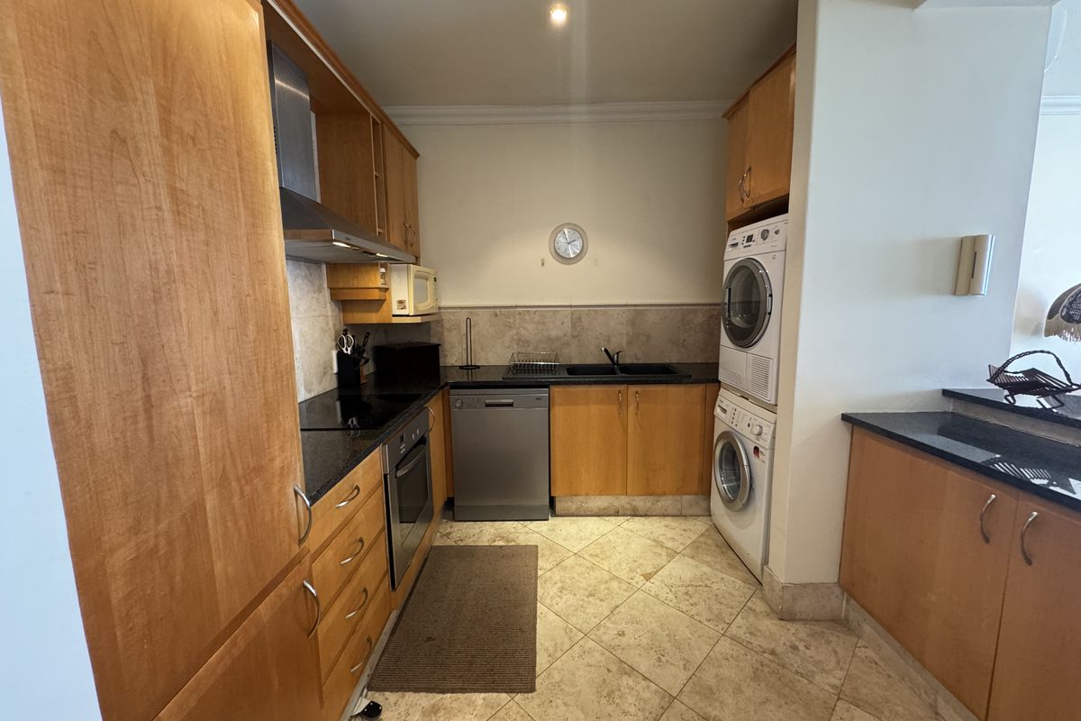 A functional kitchen featuring wooden cabinetry, granite countertops, integrated appliances, and a stacked laundry unit, finished with stone tile flooring.