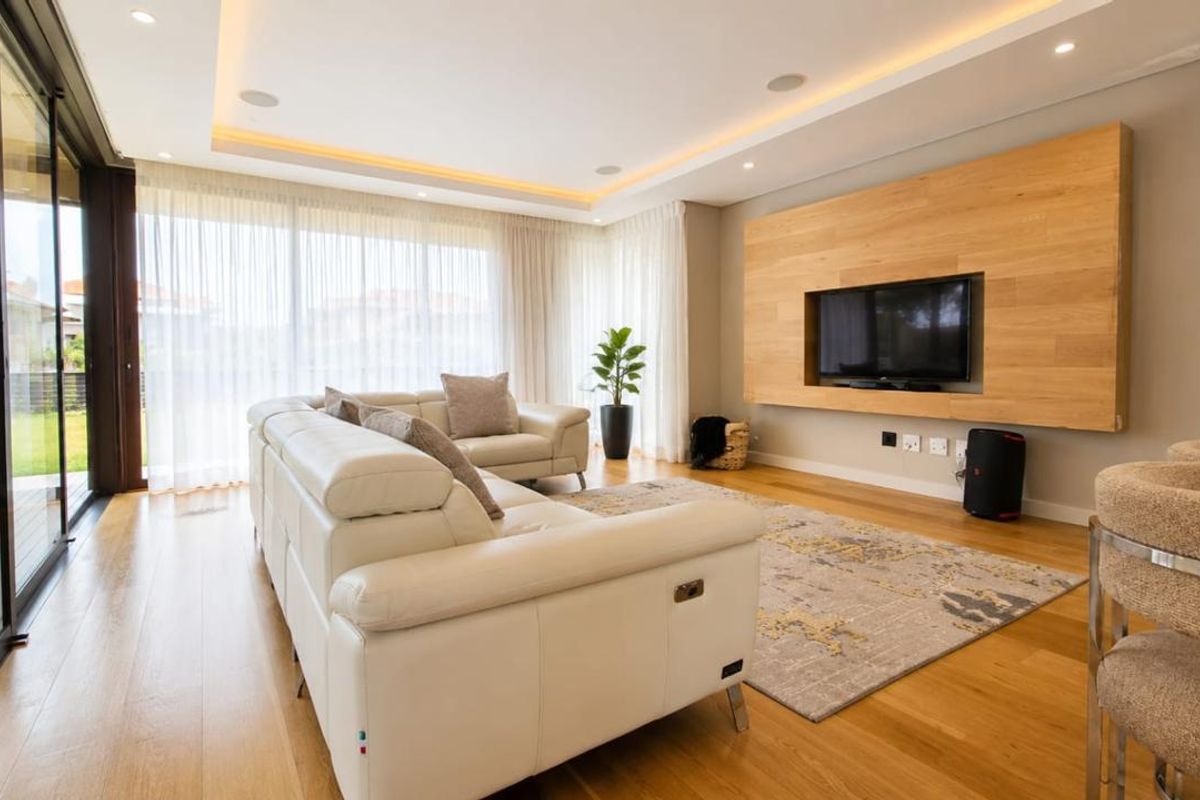 Modern living room featuring timber flooring, recessed ceiling lighting, and a custom built-in timber media wall panel with seamless access to the outdoor patio.