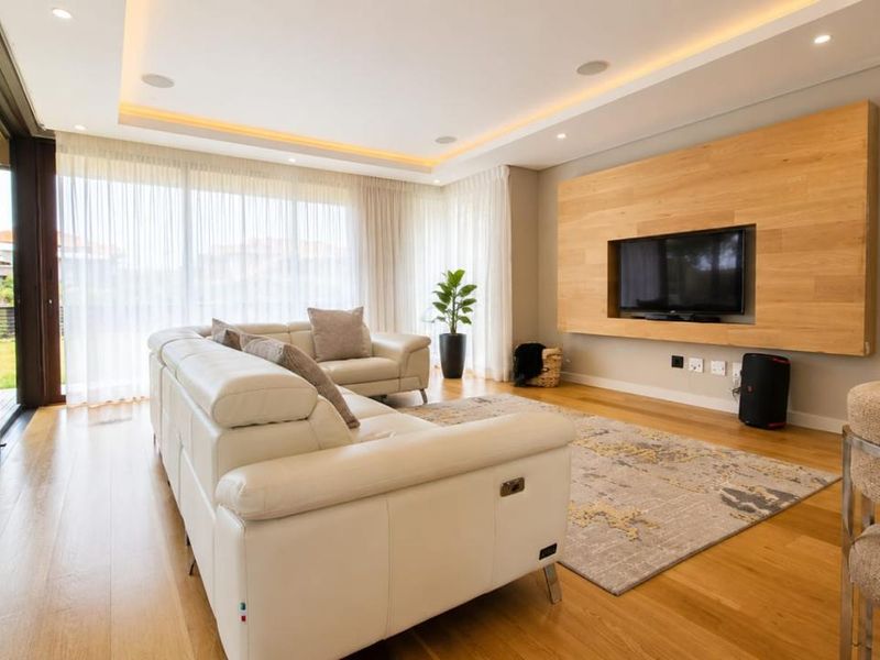 Modern living room featuring timber flooring, recessed ceiling lighting, and a custom built-in timber media wall panel with seamless access to the outdoor patio.