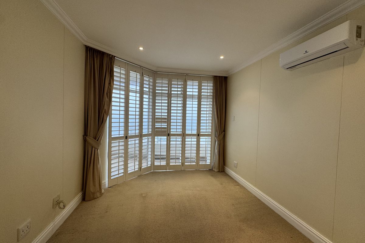 An empty living room featuring neutral walls, carpet flooring, a wall-mounted air conditioning unit, and large bay windows with plantation shutters.