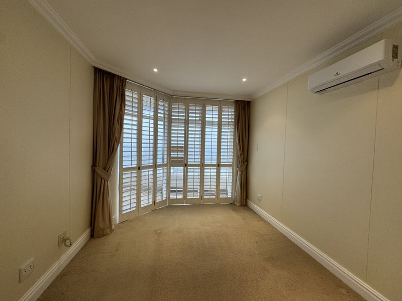 An empty living room featuring neutral walls, carpet flooring, a wall-mounted air conditioning unit, and large bay windows with plantation shutters.
